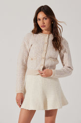 A woman is wearing a light beige, knitted cardigan with buttons down the front and two small pockets on the sides, paired with a cream-colored, textured, A-line skirt.